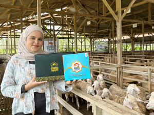 Perwakilan Oemah Aqiqah memperlihatkan dua kemasan paket aqiqah praktis dengan latar belakang kandang domba modern di Banyumas.