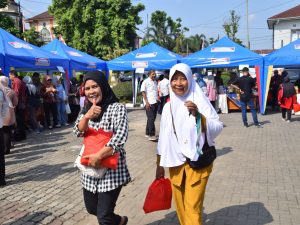 Dua warga wanita tersenyum bahagia sambil membawa kantong belanjaan sembako merah di depan deretan tenda biru Pasar Murah SARAHSIMAS DKUKMP Banyumas.