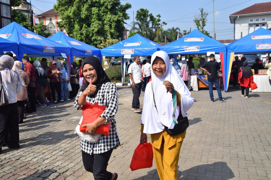 Dua warga wanita tersenyum bahagia sambil membawa kantong belanjaan sembako merah di depan deretan tenda biru Pasar Murah SARAHSIMAS DKUKMP Banyumas.