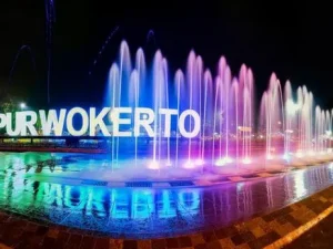 Sorotan lampu dan air mancur menari di depan landmark tulisan Purwokerto di Alun-Alun Purwokerto pada malam hari, mengilustrasikan kawasan urban Kota Purwokerto otonom.
