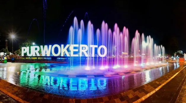 Sorotan lampu dan air mancur menari di depan landmark tulisan Purwokerto di Alun-Alun Purwokerto pada malam hari, mengilustrasikan kawasan urban Kota Purwokerto otonom.